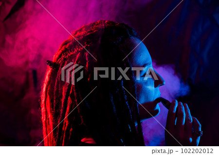 Profile of caucasian girl with dreadlocks smokes a vape in red blue light.  102202012