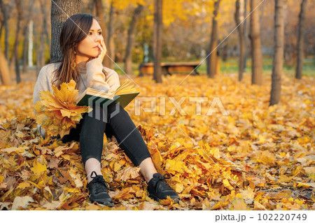 A young woman sits with a book in her hands on autumn foliage near a tree and dreams about something 102220749