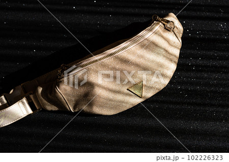 Glamorous women's waist bag in gold color on a black shiny background. Fashionable handbag with gold chain. Glamorous women's waist bag in gold color on a black shiny background. Fashionable handbag with gold chain. 102226323