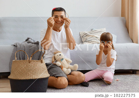 Image of dark haired male with his female child playing together at home, sitting on floor near cough in light room, making glasses with round scrunchies, having fun together, have funny expression. Image of dark haired male with his female child playing together at home, sitting on floor near cough in light room, making glasses with round scrunchies, having fun together, have funny expression. 102227575