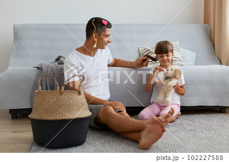 Image of Caucasian man wearing white casual style t shirt sitting on floor near sofa with child, father talking with his daughter after playing together as hairdressers. Image of Caucasian man wearing white casual style t shirt sitting on floor near sofa with child, father talking with his daughter after playing together as hairdressers. 102227588