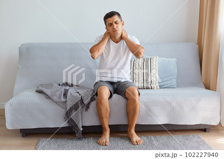 Portrait of ill unhealthy man wearing white T-shirt sitting on cough in living room, touching or massaging his painful neck, uncomfortable sleeping conditions, vertebral column injury. Portrait of ill unhealthy man wearing white T-shirt sitting on cough in living room, touching or massaging his painful neck, uncomfortable sleeping conditions, vertebral column injury. 102227940