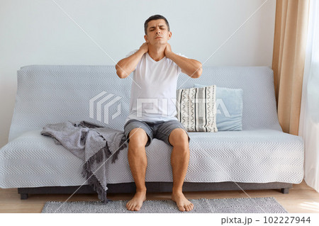 man wearing white T-shirt sitting on sofa in living room, touching neck feeling pain and numbness, worried about muscle tension, osteochondrosis, uncomfortable sleeping conditions. man wearing white T-shirt sitting on sofa in living room, touching neck feeling pain and numbness, worried about muscle tension, osteochondrosis, uncomfortable sleeping conditions. 102227944