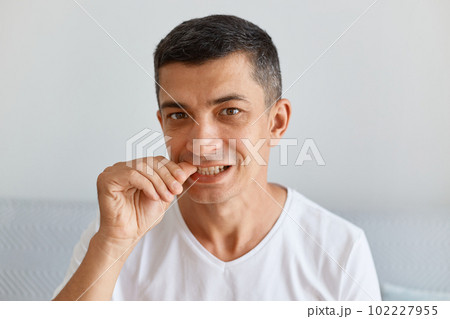 Indoor shot of Young adult Caucasian attractive brunette man wearing white shirt biting his fingernails, looking at camera, being nervous, having problems. Indoor shot of Young adult Caucasian attractive brunette man wearing white shirt biting his fingernails, looking at camera, being nervous, having problems. 102227955