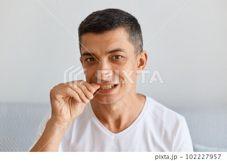 Indoor shot handsome young adult man with dark hair wearing white casual style t shirt sitting and looking at camera, biting finger nails, fells nervous. 102227957