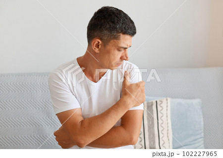 Portrait of sick young adult male with dark hair wearing white t shirt, holding his painful arm or shoulder, looking away, sitting on comfortable sofa in light living room. 102227965