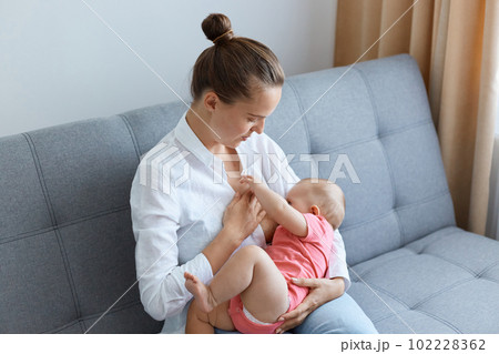 Indoor shot of attractive woman with bun hairstyle wearing white shirt sitting on sofa with infant baby, young mother breastfeeding her cute toddler daughter. Indoor shot of attractive woman with bun hairstyle wearing white shirt sitting on sofa with infant baby, young mother breastfeeding her cute toddler daughter. 102228362