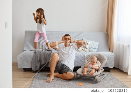 Indoor shot of young adult man sitting on floor near sofa and covering ears with palms, being tired of loud daughter jumping on floor and scream, wearing white t shirt and jeans short. 102228813