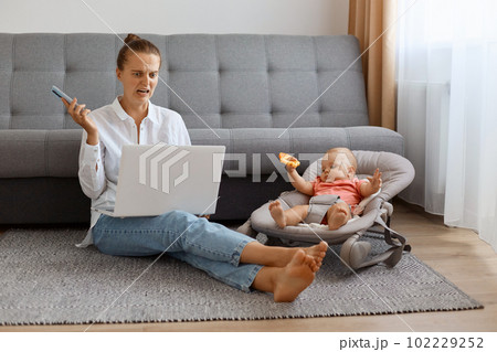 Caucasian woman, young mother working on laptop, sitting on floor with her daughter in rocking chair, looking at laptop display with disguast, sees forbidden content. Caucasian woman, young mother working on laptop, sitting on floor with her daughter in rocking chair, looking at laptop display with disguast, sees forbidden content. 102229252