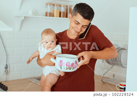 Indoor shot of smiling dark haired father talking via mobile phone and holding baby daughter and pot in hands, man taking care of his kid, being in paternity leave. Indoor shot of smiling dark haired father talking via mobile phone and holding baby daughter and pot in hands, man taking care of his kid, being in paternity leave. 102229324
