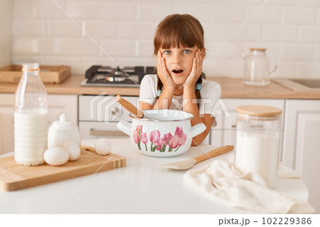 Portrait of surprised amazed little cute girl with dark hair and braids wearing white t shirt cooking in light kitchen, looking at camera with open mouth, forgot recipe of her favorite cakes. 102229386
