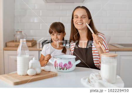 Beautiful young adult mom and her cute little daughter playing and smiling while baking in kitchen at home, looking at camera with happy smiling. 102229450