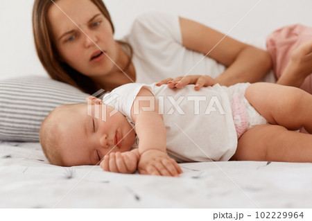 Portrait of scared worried mother looking at her little sleeping daughter, touching baby, female with dark hair wearing white casual style t shirt, motherhood. 102229964