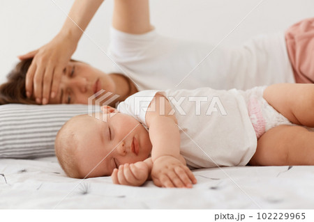 Tired exhausted female wearing white casual style t shirt lying near sleeping baby, keeping palm on forehead, lying with child, having sleepless night, motherhood. Tired exhausted female wearing white casual style t shirt lying near sleeping baby, keeping palm on forehead, lying with child, having sleepless night, motherhood. 102229965