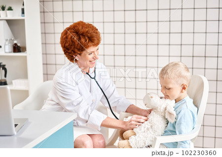 Female Pediatrician listening to breathing in the lungs and heartbeat with stethoscope. Portrait of adorable little boy visiting doctor. Female Pediatrician listening to breathing in the lungs and heartbeat with stethoscope. Portrait of adorable little boy visiting doctor. 102230360