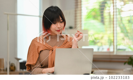 Charming young asian woman in stylish clothes working on laptop computer at modern home office 102233183