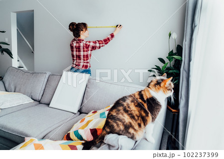 Young woman doing measuring with a measure tape on the wall. Girl wants to put a picture on the wall at home. Housekeeping work. Doing repair herself. DIY, gender equality in work concept. Young woman doing measuring with a measure tape on the wall. Girl wants to put a picture on the wall at home. Housekeeping work. Doing repair herself. DIY, gender equality in work concept. 102237399