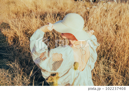 Tender back view woman in light airy dress and straw hat holding blade of grass and enjoying the moment in a high grass meadow at sunset. summer evening in nature. Calm and harmony. Vacation. 102237408