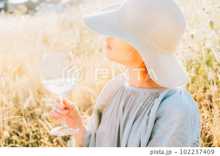 Closeup unrecognizable woman in light airy dress and straw hat holding wine glass looking at sunset in a high grass meadow. Enjoy summer evening on nature. Calm and harmony. Vacation. Selective focus. 102237409