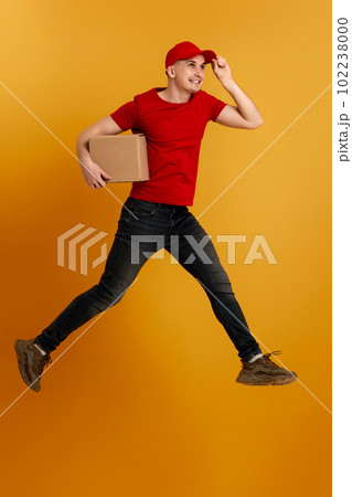 Delivery man in red cap, t-shirt holding cardboard box and jumping Delivery man in red cap, t-shirt holding cardboard box and jumping 102238000