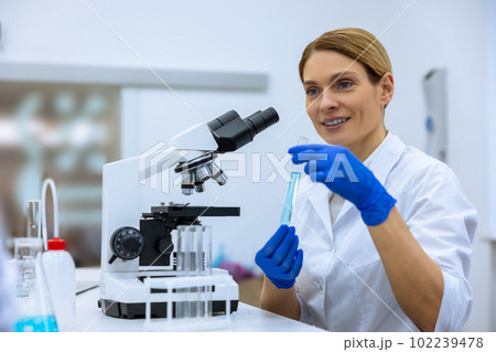 Scientist working in scientific laboratory, holding test tube with blue liquid, using microscope Scientist working in scientific laboratory, holding test tube with blue liquid, using microscope 102239478