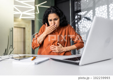 Businesswoman has severe nausea, latin american woman at work sick, working sitting at table with laptop inside office Businesswoman has severe nausea, latin american woman at work sick, working sitting at table with laptop inside office 102239993