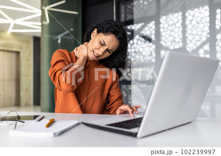 Overtired business woman working inside office with laptop, Hispanic female worker has severe neck pain, massaging muscles with hand Overtired business woman working inside office with laptop, Hispanic female worker has severe neck pain, massaging muscles with hand 102239997