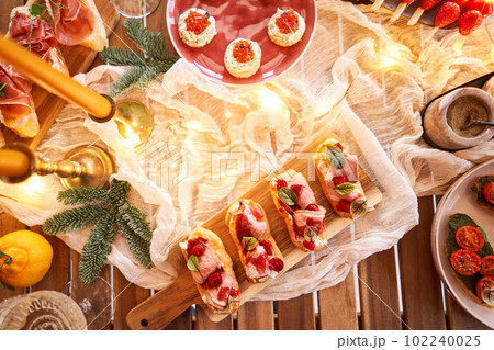 Christmas dinner feast. A small table is served with snacks, bruschettas, and canapes. A decorated dining table with champagne glasses, candles and christmas tree an garland in background Christmas dinner feast. A small table is served with snacks, bruschettas, and canapes. A decorated dining table with champagne glasses, candles and christmas tree an garland in background 102240025