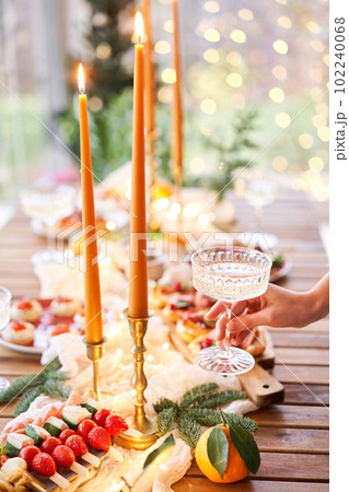 Hand with a glass of champagne in the foreground. A small table is served with snacks, bruschettas, and canapes. A decorated dining table with champagne glasses, candles and christmas tree an garland 102240068