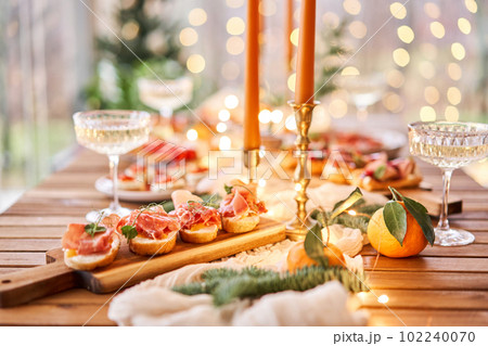 Christmas dinner feast. A small table is served with snacks, bruschettas, and canapes. A decorated dining table with champagne glasses, candles and christmas tree an garland in background Christmas dinner feast. A small table is served with snacks, bruschettas, and canapes. A decorated dining table with champagne glasses, candles and christmas tree an garland in background 102240070