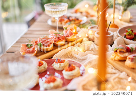 Christmas dinner feast. A small table is served with snacks, bruschettas, and canapes. A decorated dining table with champagne glasses, candles and christmas tree an garland in background Christmas dinner feast. A small table is served with snacks, bruschettas, and canapes. A decorated dining table with champagne glasses, candles and christmas tree an garland in background 102240071