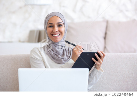 Close-up photo. Muslim young woman in hijab freelancer work from home online. Sitting on the sofa with a laptop and a folder with documents. He writes, fills in with a pen, smiles at the camera. Close-up photo. Muslim young woman in hijab freelancer work from home online. Sitting on the sofa with a laptop and a folder with documents. He writes, fills in with a pen, smiles at the camera. 102240464