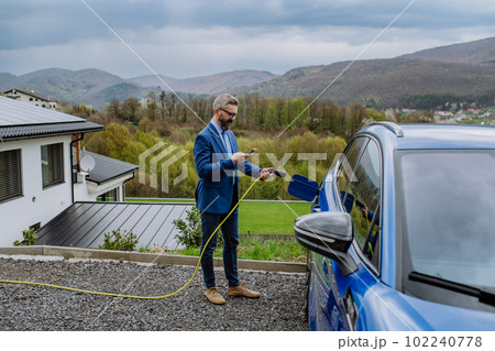 Mature businessman holding power supply cable,scrolling his phone and charging his electric car. Mature businessman holding power supply cable,scrolling his phone and charging his electric car. 102240778