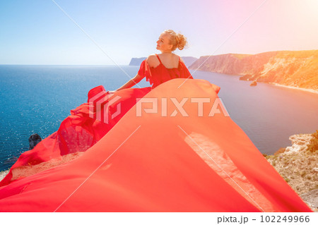 Blonde with long hair on a sunny seashore in a red flowing dress, back view, silk fabric waving in the wind. Against the backdrop of the blue sky and mountains on the seashore. 102249966