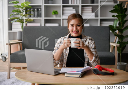 Attractive Asian female freelancer sits at a coffee table in her living room with a coffee cup Attractive Asian female freelancer sits at a coffee table in her living room with a coffee cup 102251019