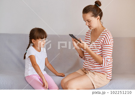 Professional female physiotherapist working with small child girl, sitting on sofa with mobile phone in hands, finds new online lesson in her gadget for training. Professional female physiotherapist working with small child girl, sitting on sofa with mobile phone in hands, finds new online lesson in her gadget for training. 102251210