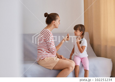 Woman wearing striped shirt and shorts sitting on sofa with little girl, articulation exercised at lesson at home, mother or teacher and female kid learn pronouncing sounds together. Woman wearing striped shirt and shorts sitting on sofa with little girl, articulation exercised at lesson at home, mother or teacher and female kid learn pronouncing sounds together. 102251234