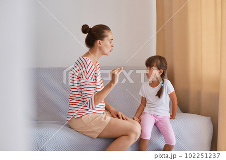 Side view of woman with bun hairstyle wearing striped shirt and shorts sitting on sofa with daughter, preschooler girl practicing correct pronunciation with a female speech therapist. 102251237
