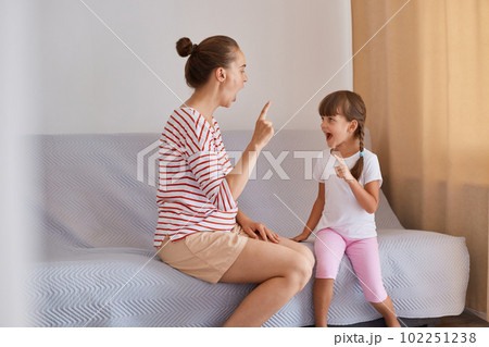 Indoor shot of hard working woman speech therapist working with little girl at home, female wearing striped shirt and shorts sitting on sofa with child. 102251238