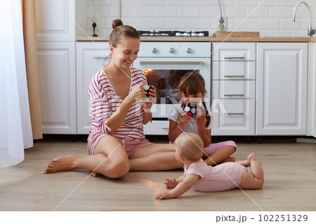 Portrait of attractive female with hair ban wearing striped casual shirt sitting on floor in kitchen with cup in hands, spending time with her little daughters. 102251329