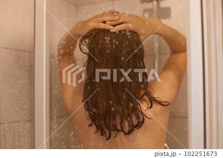 Back side photo of dark haired woman with naked body takes shower behind glass door in bathroom, enjoys relaxation in bathroom, has healthy pure skin. 102251673