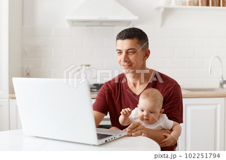 Positive handsome man with dark hair wearing burgundy casual t shirt, looking a notebook screen, working on laptop while babysitting, posing in white kitchen. Positive handsome man with dark hair wearing burgundy casual t shirt, looking a notebook screen, working on laptop while babysitting, posing in white kitchen. 102251794