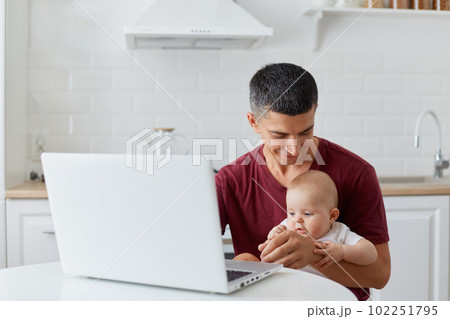 Indoor shot of young adult father wearing maroon casual t shirt sitting with little daughter or son in front of notebook, looking at baby with great love, freelancer work while taking care of child. 102251795