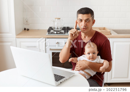 Smart brunette male wearing maroon casual style t shirt sitting at table in kitchen with his infant daughter, having clever idea, raising his finger up. Smart brunette male wearing maroon casual style t shirt sitting at table in kitchen with his infant daughter, having clever idea, raising his finger up. 102251796