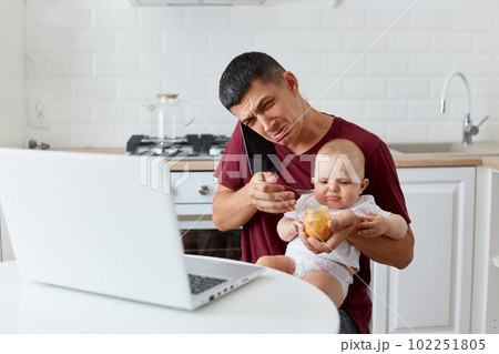 Portrait of sad upset man wearing maroon casual t shirt sitting with little daughter or son on knees, talking phone and crying, needs work and take care of baby, has no time. Portrait of sad upset man wearing maroon casual t shirt sitting with little daughter or son on knees, talking phone and crying, needs work and take care of baby, has no time. 102251805
