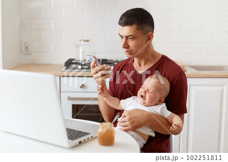 Shocked brunette male wearing maroon casual style t shirt sitting at table in kitchen with his infant daughter, looking at smartphone display with scared expression. Shocked brunette male wearing maroon casual style t shirt sitting at table in kitchen with his infant daughter, looking at smartphone display with scared expression. 102251811