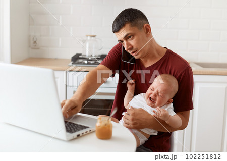 Indoor shot of brunette male wearing maroon casual style t shirt sitting at table in kitchen with his infant daughter, talking via cell phone while working online on laptop. Indoor shot of brunette male wearing maroon casual style t shirt sitting at table in kitchen with his infant daughter, talking via cell phone while working online on laptop. 102251812