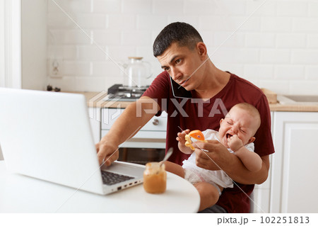 Portrait of busy brunette male freelancer wearing maroon casual style t shirt sitting at table in kitchen with his infant daughter and talking via cell phone with customer. 102251813