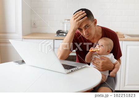 Exhausted attractive brunette man freelancer wearing casual style maroon t shirt, working and looking after his little baby daughter, keeping hand on his forehead. 102251823