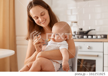 Dark haired female feeding her little daughter with fruit or vegetable puree, looking at baby with charming smile, holding spoon in hand, looking at toddler girl. Dark haired female feeding her little daughter with fruit or vegetable puree, looking at baby with charming smile, holding spoon in hand, looking at toddler girl. 102251925